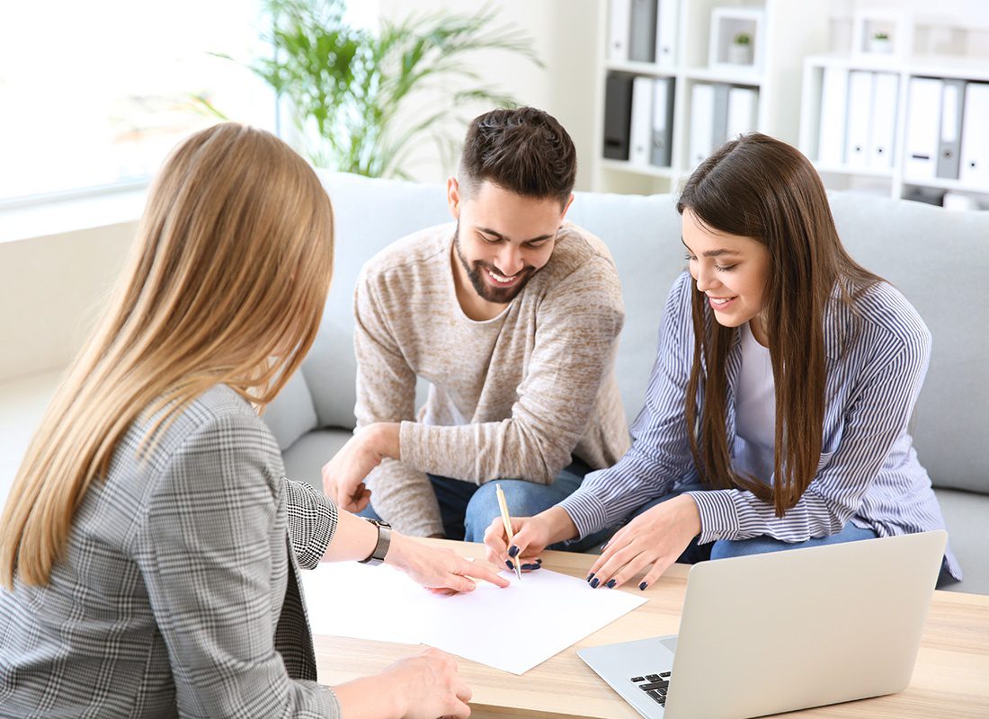 About Our Agency - Happy Couple Sit Together on a Sofa Across From Their Agent as They Sign Some Documents